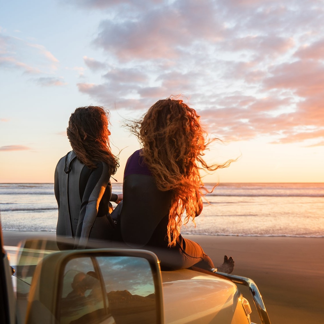 girls sitting on car bonnet looking at beach at sunset